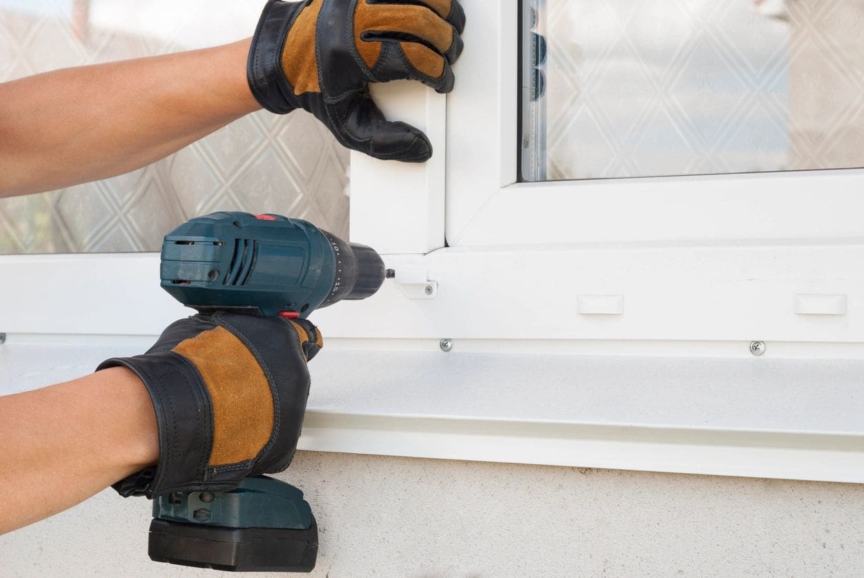 A person wearing gloves holding a drill near a window.